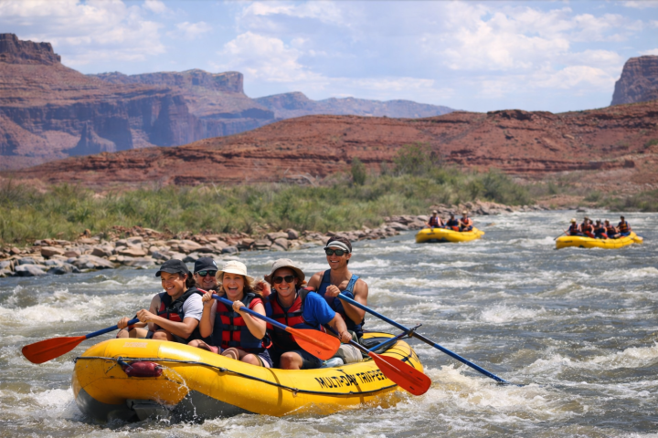 Group of people whitewater rafting on a river with desert landscape and mountains in the background.