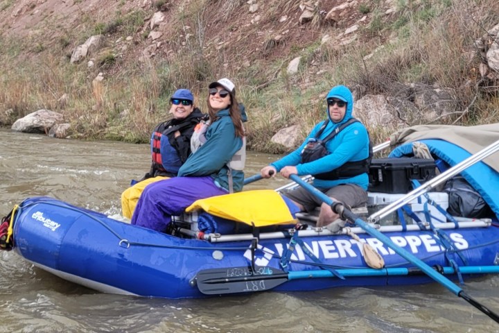 Three people rafting on a river in a blue inflatable boat near a rocky bank.