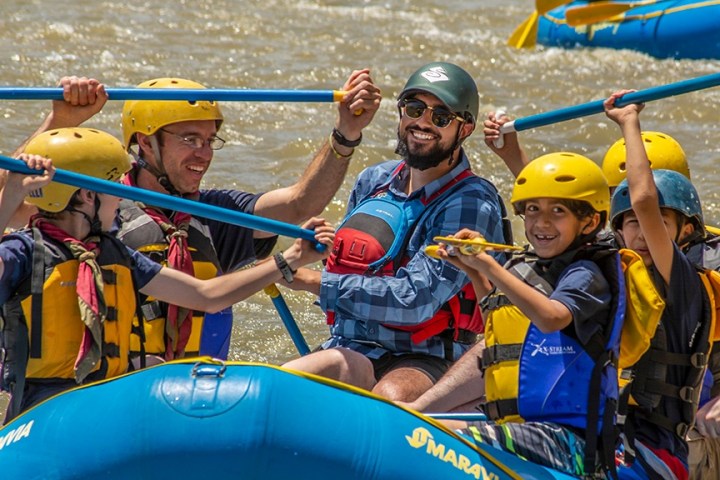 Group of people rafting on a river, wearing helmets and life jackets, smiling and holding paddles.