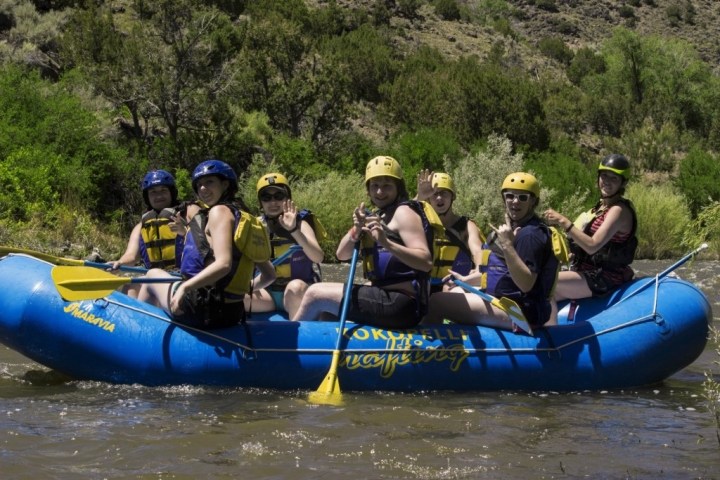 Group of people in blue raft with paddles, wearing helmets and life vests, on a river.