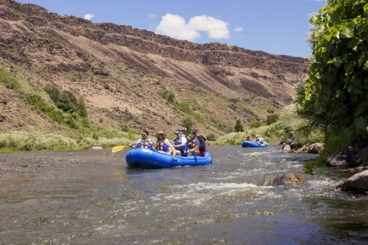 People in blue rafts rafting on a river with rocky hills and green shrubs in the background.