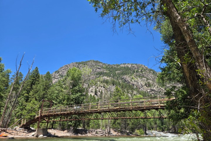 Wooden footbridge over a river, surrounded by trees and a mountain backdrop.
