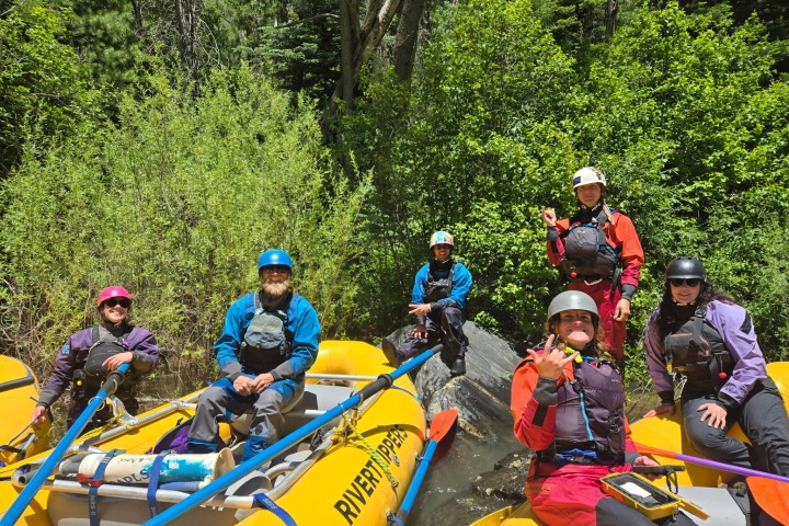 Group of six people in rafting gear sitting on yellow inflatable rafts near lush greenery.