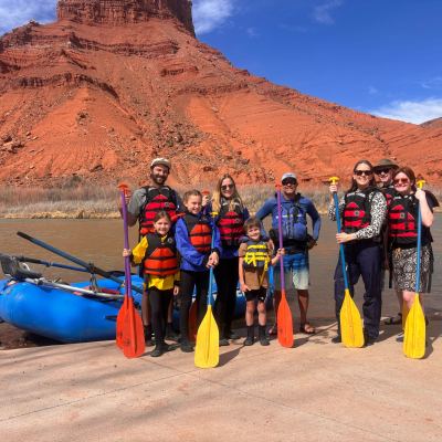 Group in life jackets with paddles by a river and red rock cliff.