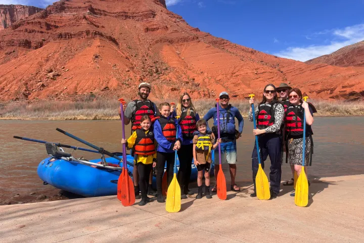 Group in life jackets with paddles by a river and red rock cliff.