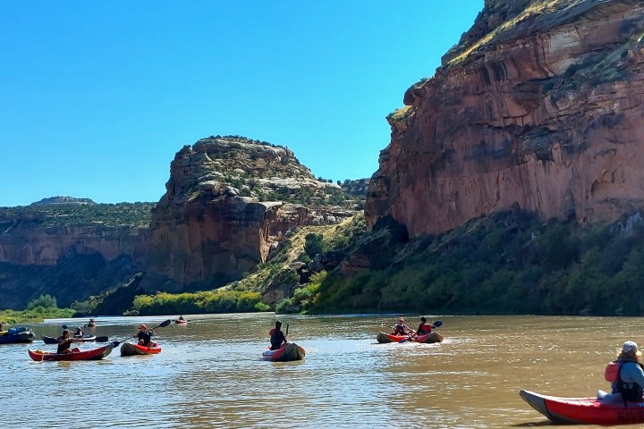 People kayaking on river with cliffs under clear blue sky.
